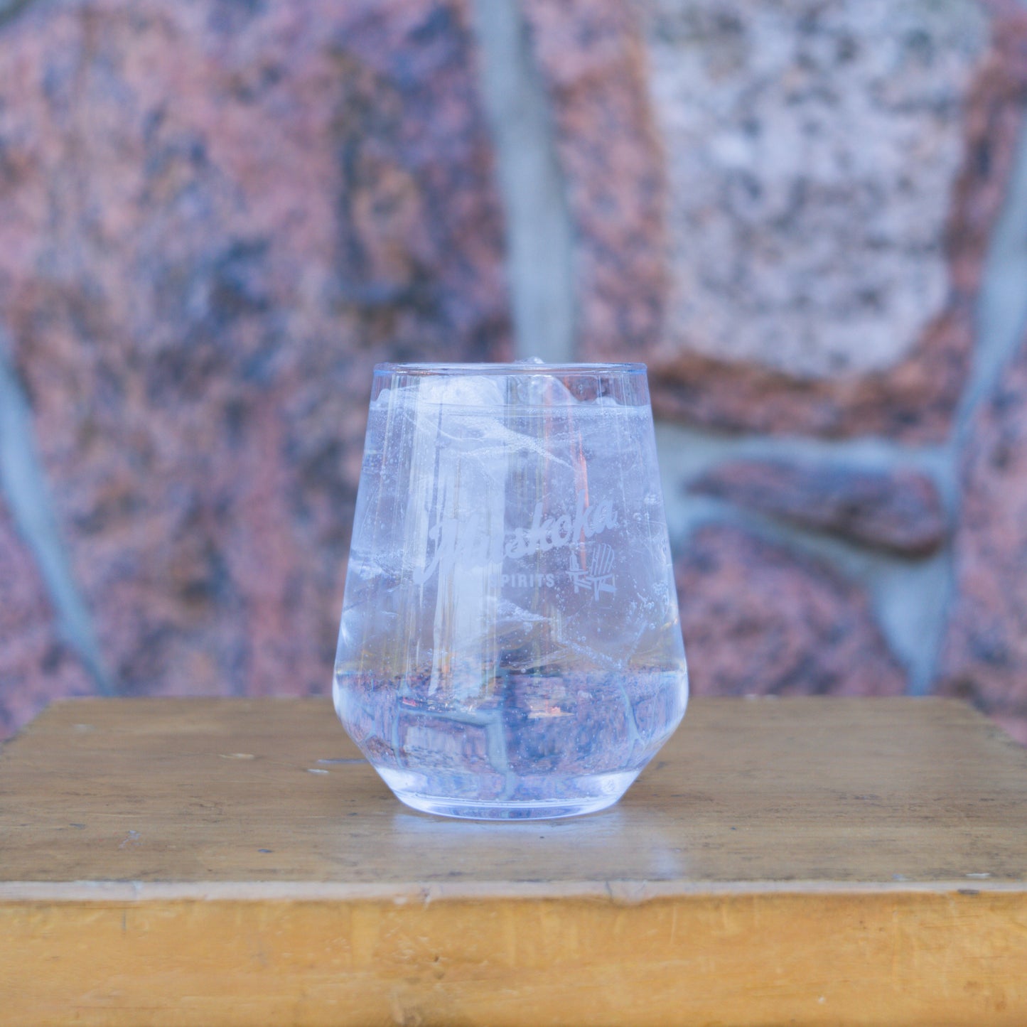 A clear stemless cocktail glass filled with ice, displayed on a wooden surface with a rock wall in the background.